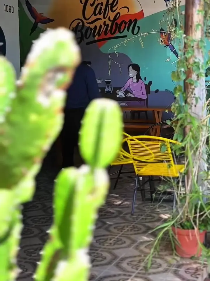 Varanda do Bourbon com o mural colorido 'Café Bourbon', lanternas azuis, cadeiras amarelas e plantas.