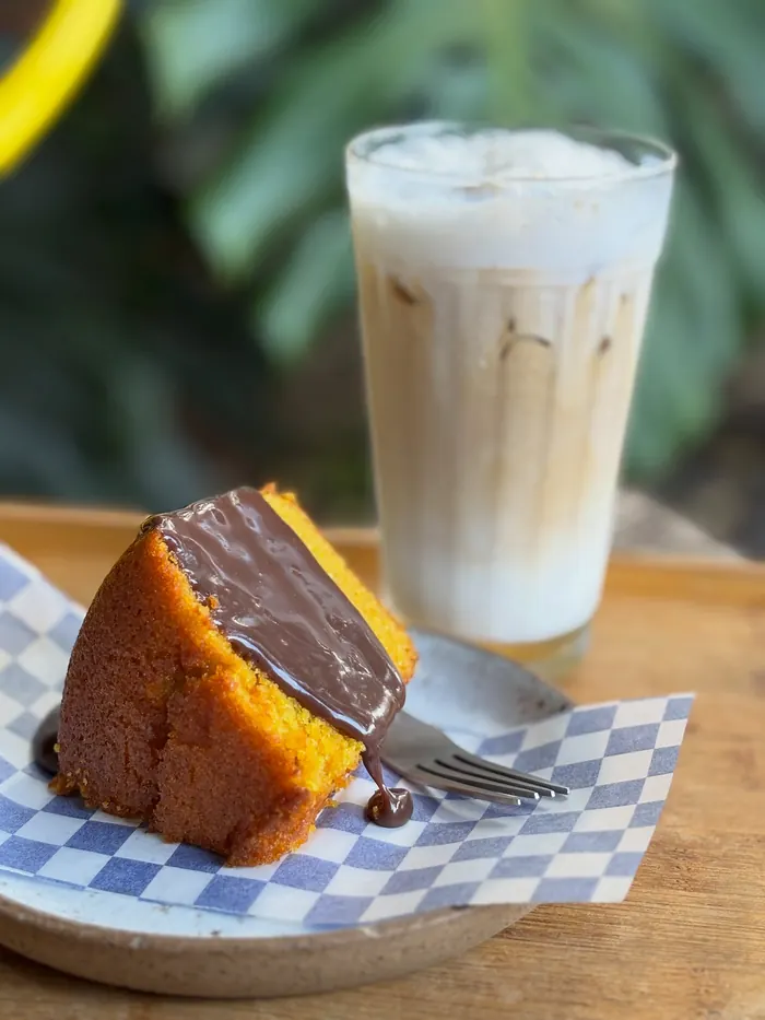Fatia de bolo de cenoura com cobertura de chocolate e um latte gelado ao fundo.