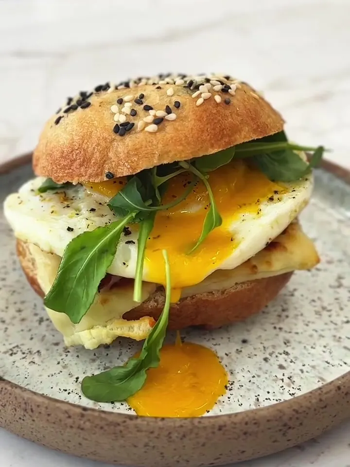Bagel com gergelim recheado com queijo coalho, bacon, ovo de gema mole e rúcula, num prato de cerâmica.