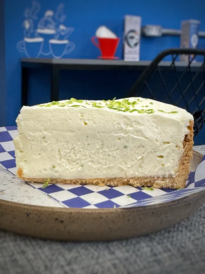 Fatia alta de torta de limão com raspas da fruta, sobre papel xadrez azul.