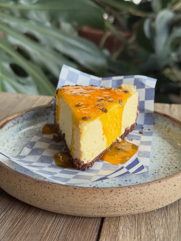 Fatia de torta de maracujá com calda da fruta sobre papel xadrez azul, num prato de cerâmica.