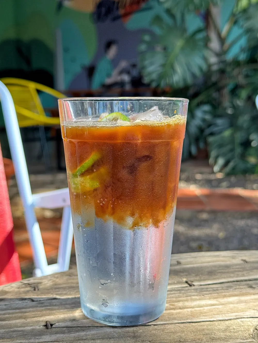 Café gelado com limão num copo de vidro, na varanda do Bourbon, com o mural colorido, plantas e uma cadeira amarela ao fundo.
